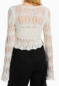 Biały top wykonany na szydełku, ozdobiony kwiatowym wzorem koronki, z długimi rękawami i krótkim fasonem, zestawiony z czarnymi, wysokim stanem spodniami.