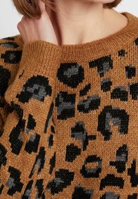 Brązowy sweter w czarno-szare wężowe wzory, prążkowany dekolt i mankiety, miękka tekstura, zbliżenie na szczegóły rękawa i dekoltu.