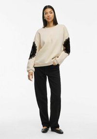 Beige sweatshirt with black lace detailing on sleeves, relaxed fit. Paired with black wide-leg pants and flat shoes. Smooth texture.