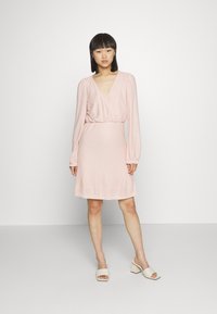 Vestido rosa claro com mangas longas, decote em v e cintura franzida. Tecido texturizado com um padrão subtil. Combinado com sandálias de salto bloco brancas.