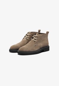 Botas de ante marrón hasta el tobillo con puntera redonda, diseño de cordones y suelas de goma negra contrastantes; con una tira en la parte superior para facilitar su colocación.