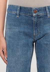 Personne portant un jean en denim bleu et un haut beige à côtes, montrant la taille et la main reposant sur le côté contre un fond uni.