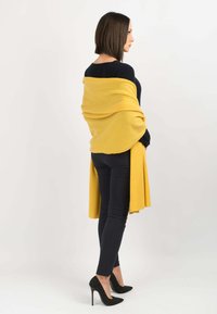 Sciarpa di lana gialla drapeata su una maglia blu navy, indossata con pantaloni neri attillati e tacchi a pois. La superficie liscia e la forma allungata accentuano la silhouette.