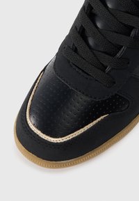 Czarne sneakersy z gładkim skórzanym wierzchem, z perforowanymi sekcjami. Złote akcenty wzdłuż palców oraz teksturowana gumowa podeszwa. Czarne sznurowadła.