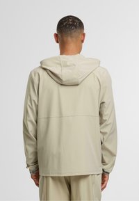 Béžová lehká bunda s kapucí s hladkým povrchem, disponující horizontálním švem na zádech a elastickými manžetami.