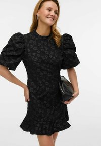 Črna obleka z nabranimi rokavi, cvetličnim vzorcem z reliefno teksturo, oprijetim trupom in razširjenim robom. Oblečena s majhno črno torbico.