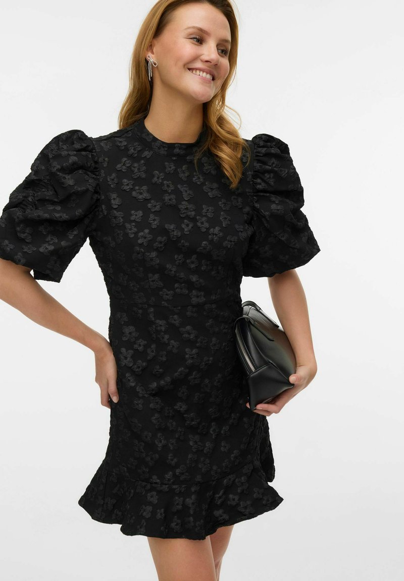 Črna obleka z nabranimi rokavi, cvetličnim vzorcem z reliefno teksturo, oprijetim trupom in razširjenim robom. Oblečena s majhno črno torbico.