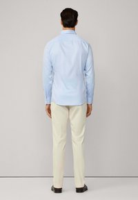 Muškarac stoji okrenut prema zidu, obučen u svijetloplavu košulju dugih rukava, krem boje hlače i tamno smeđe cipele.