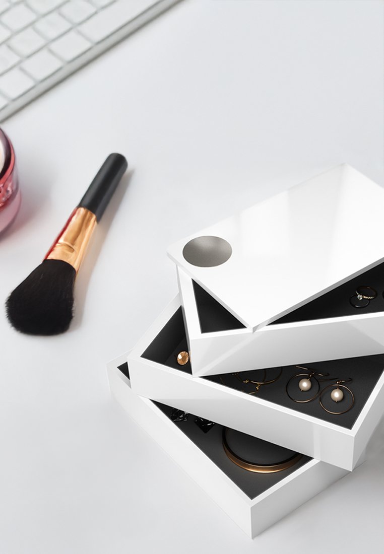 Organizador de joyas geométrico blanco con tres bandejas apiladas, cada una con un interior negro. Incluye varios artículos de joyería visibles en el interior.
