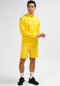 Gul atletisk träningsoverall med zip-up jacka och shorts. Har svarta axelränder. Bärs med svarta sneakers och vita strumpor.