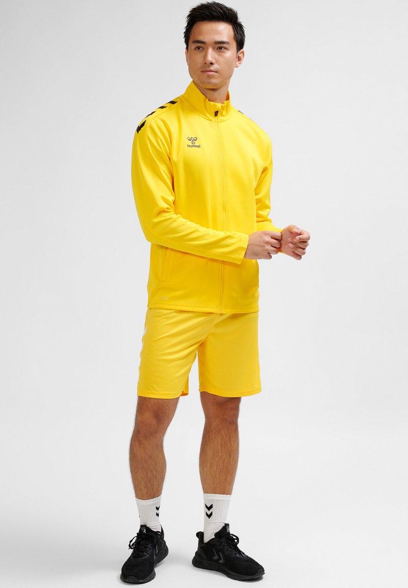 Gul atletisk träningsoverall med zip-up jacka och shorts. Har svarta axelränder. Bärs med svarta sneakers och vita strumpor.