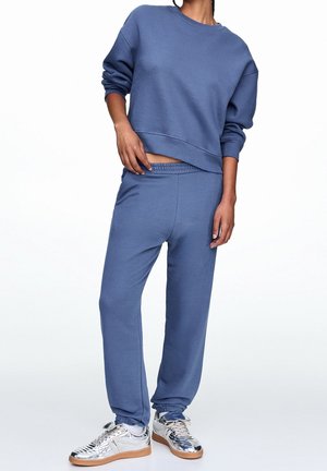 Personne portant un sweat-shirt bleu à manches longues et un pantalon de survêtement assorti avec des baskets métalliques argentées, debout devant un fond blanc.
