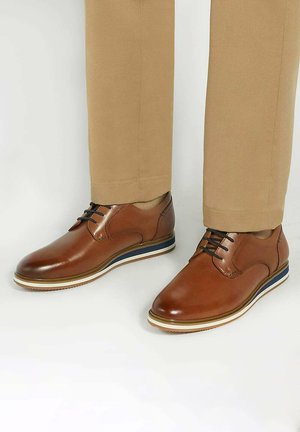 Bruine leren dresschoenen met ronde neuzen, zwarte veters en een gelaagde zool met blauwe en witte accenten, gecombineerd met beige broek.