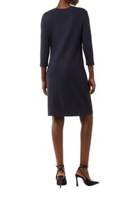 Granatowa dzianinowa sukienka z rękawami o długości trzech ćwiartek i bocznymi rozcięciami, z okrągłym dekoltem i luźnym krojem. Zestawiona z czarnymi butami na wysokim obcasie.