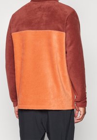 Sudadera de forro polar en óxido y naranja, con un acabado texturizado y suave. Presenta un cuello alto y un diseño de bloques de color contrastantes en la parte posterior.
