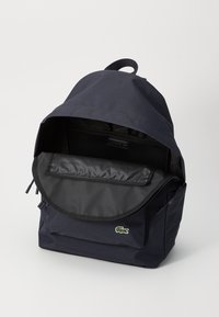 Marineblauwe rugzak met een ronde bovenkant, gemaakt van stof, met een voorzak en een logo. Het interieur heeft een gaasvak en een gevoerd compartiment.
