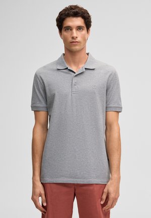 Jonge man met kort krullend haar, gekleed in een grijs, kort mouwig poloshirt en roodbruine broek, staand tegen een effen achtergrond.