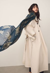 Trench beige con grandi bottoni neri e tasche, indossato sopra una sciarpa blu testurizzata con motivi animali in grigio e accenti dorati.