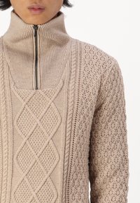 Pull en tricot beige avec un col montant et une fermeture éclair devant. Présente des motifs texturés, y compris un motif en diamant et des accents en tricot torsadé.