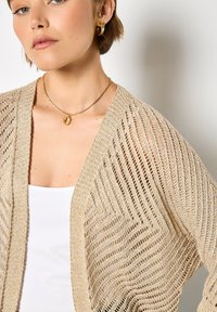 Jasnobeżowy sweter z dzianiny, zapinany na przodzie, z geometrycznym wzorem i luźnymi rękawami. Noszony na białym topie na ramiączkach.