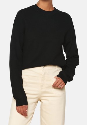 Personne portant un pull noir à manches longues et un pantalon taille haute blanc cassé avec une main dans la poche.