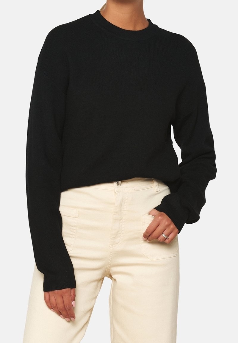 Personne portant un pull noir à manches longues et un pantalon taille haute blanc cassé avec une main dans la poche.