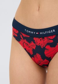 Μπικίνι σλιπ Tommy Hilfiger με ναυτικό μπλε βάση και μεγάλα κόκκινα ανθισμένα μοτίβα, ζώνη μέσης σε αντίθεση με λεπτομέρειες λογοτύπου και απαλή υφή υφάσματος.