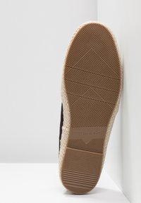 Sort fodtøj med en vævet jute-lignende sål. Har en tekstureret gummibund med et geometrisk mønster og trykt brandnavn.