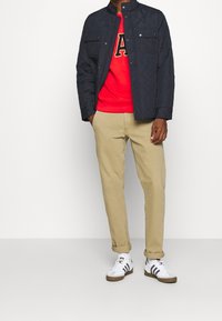 Uomo che indossa una giacca nera trapuntata, una felpa rossa con stampa di lettere, pantaloni beige arrotolati e scarpe da ginnastica bianche con strisce nere.