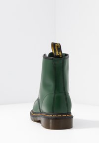 Botte unique en cuir vert avec semelle noire, couture jaune, et languette de tirage noire avec texte jaune à l'arrière, vue de dos.