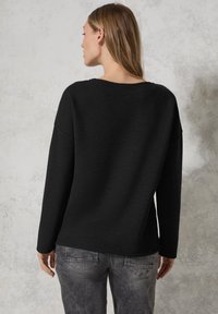 Schwarzer strukturierter Pullover mit lockerer Passform und fallenden Schultern, mit einem runden Halsausschnitt. Kombiniert mit grauen Jeans, von hinten betrachtet.