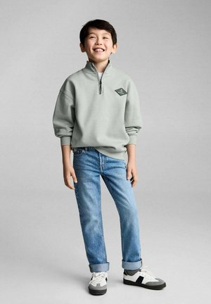 Lichtgrijze, rits-hoodie met een logo op de borst, gecombineerd met blauwe spijkerbroek en witte sneakers met grijze accenten. Simpele en casual outfit.