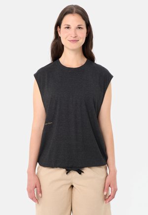 Junge Frau mit langen braunen Haaren, die ein dunkelgraues ärmelloses Oberteil und beige Hosen trägt, steht vor einem schlichten hellen Hintergrund.