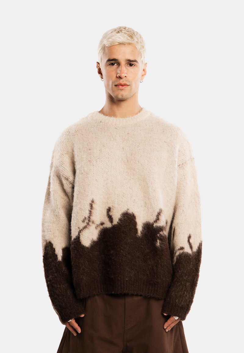 Jeune personne aux cheveux blonds courts portant un pull pelucheux beige et marron foncé oversized et un pantalon marron, debout devant un fond uni.