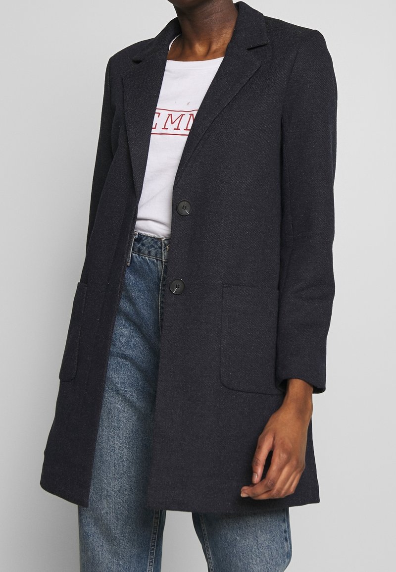 Cappotto in misto lana blu navy, con chiusura a un petto, due tasche frontali, rever strutturati e maniche a tre quarti, abbinato a una t-shirt bianca e jeans.