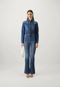 Γυναίκα ντυμένη με εφαρμοστό μπλε τζάκετ από τζιν και καμπάνα τζιν παντελόνι, κρατώντας μια λευκή τσάντα, με μακριά ίσια μαλλιά και χρυσά σκουλαρίκια.