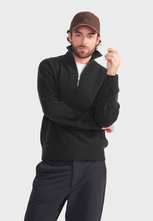 Zwarte geribbelde gebreide pullover met een halve ritssluiting, gecombineerd met donkere broeken. De textuur is dik, wat zorgt voor een comfortabele uitstraling.
