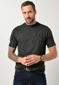 Camiseta negra de manga corta con texto en relieve "SALT LAKE", diseño de cuello redondo, combinada con jeans denim oscuros y un reloj negro.