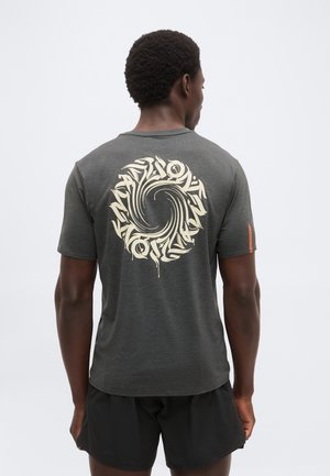 Mand iført mørkegrå t-shirt med cirkulært abstrakt cremefarvet mønster på ryggen og sorte shorts, stående mod en ensfarvet lys baggrund.