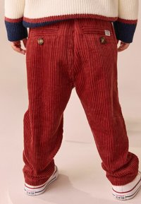 Rote Cordhosen mit gerippter Textur, ausgestattet mit Gesäßtaschen, Knöpfen und einem lässigen Schnitt, kombiniert mit weißen Turnschuhen.