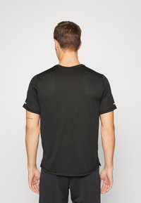 Man som bär en enkel svart kortärmad träningsskjorta och svarta shorts, sedd bakifrån mot en enfärgad ljus bakgrund.