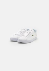 Hvite kanvas sneakers med snøring, gummisåler og en liten grønn logoaktig detalj på siden. Glatt tekstur og minimalistisk form.