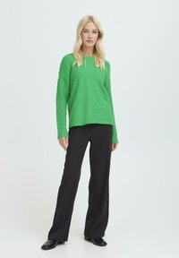 Žalias megztinis su laisvu siluetu ir įbrėžtu kaklu, derinamas su juodomis plačiomis kelnių ir juodais batais, šviesiame fone.