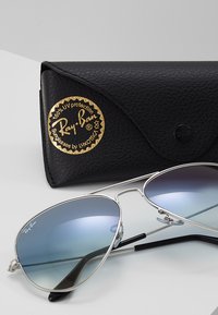 Ray-Ban sunglasses with silver metal frame, blue gradient lenses. Accompanied by a black textured case featuring a gold logo badge.