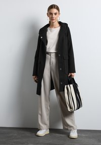 Abrigo negro con capucha y botones plateados, llevado sobre una blusa clara y un pantalón beige. El modelo sostiene un bolso de mano a rayas en negro y crema.