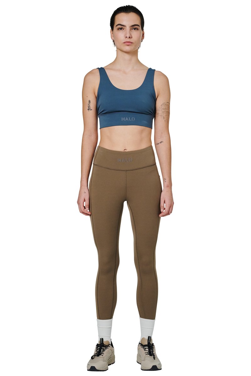 Kvinde stående iført blå sports-bh og olivengrønne leggings med hvide sokker og beige sneakers mod en hvid baggrund.