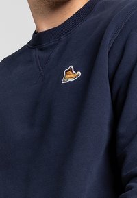 Marineblaue Sweatshirt mit strukturiertem Finish, das auf der linken Brustseite ein kleines gesticktes Logo eines braunen Stiefels aufweist.