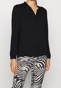 Blouse noire à manches longues avec un décolleté en V et un détail de surpiqûre subtile, associée à un legging rayé noir et blanc avec un motif ondulé.