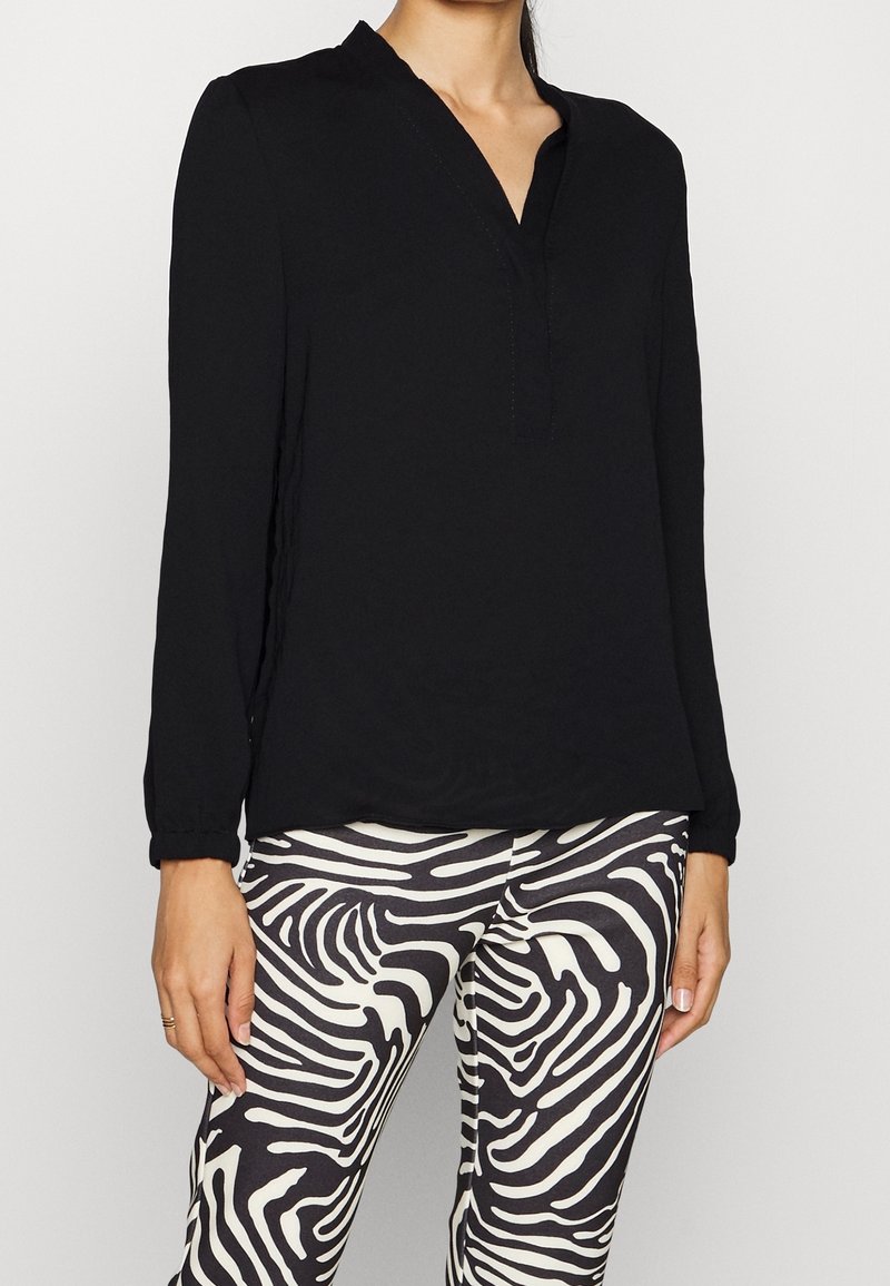 Blouse noire à manches longues avec un décolleté en V et un détail de surpiqûre subtile, associée à un legging rayé noir et blanc avec un motif ondulé.