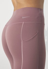 Leggings de cintura alta en tela malva, con un bolsillo lateral discreto, costuras en contraste y un pequeño logo gris cerca de la cinturilla.
